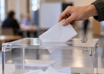 Una mano introduce un sobre de votación en una urna electoral transparente en un colegio electoral