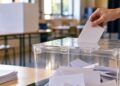 Mano depositando un sobre blanco en una urna electoral transparente en un aula, sin rostros visibles