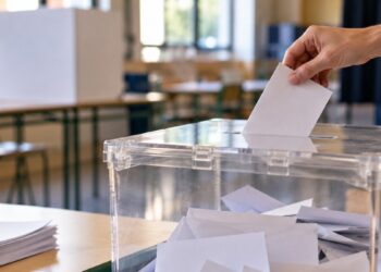 Mano depositando un sobre blanco en una urna electoral transparente en un aula, sin rostros visibles